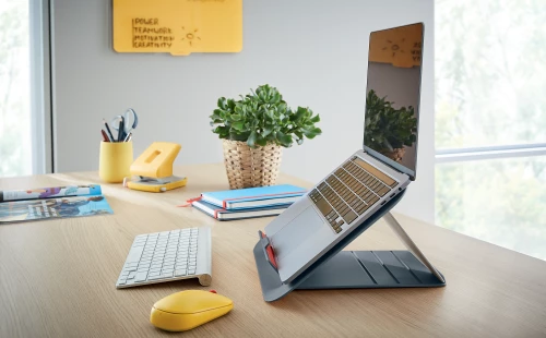szara podstawka z laptopem stojąca na biurku, przed białą klawiaturą i żółtą myszką