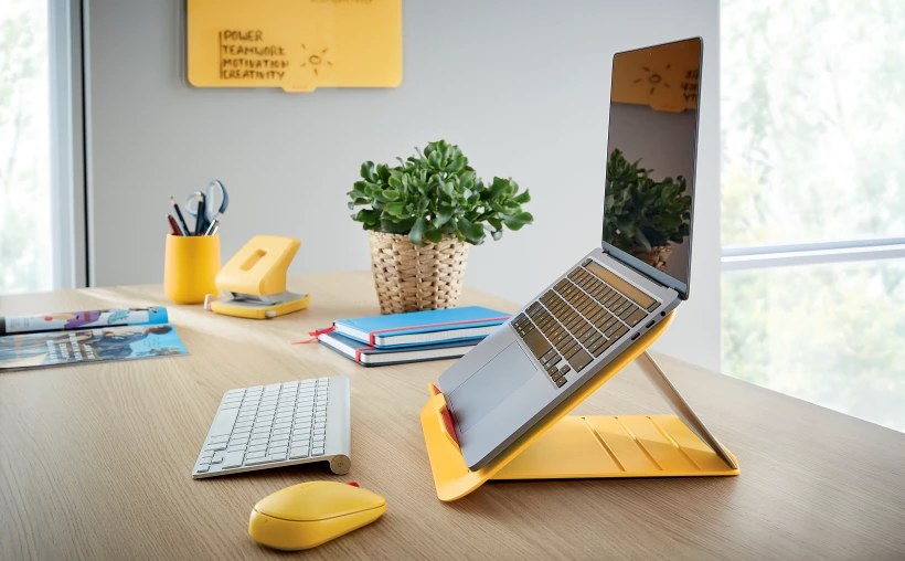 żółta podstawka z laptopem stojąca na biurku, przed białą klawiaturą i żółtą myszką