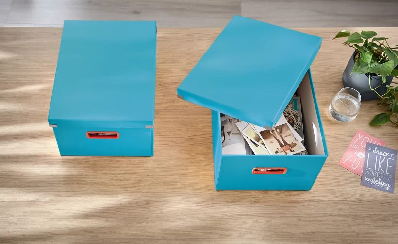 niebieskie pudełko z otwartą pokrywą, z umieszczonymi wewnątrz zdjęciami, położone na biurku w towarzystwie drugiego niebieskiego pojemnika z zamkniętą pokrywą