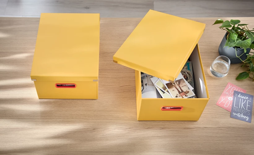 żółte pudełko z otwartą pokrywą, z umieszczonymi wewnątrz zdjęciami, położone na biurku obok drugiego żółtego pojemnika z zamkniętą pokrywą