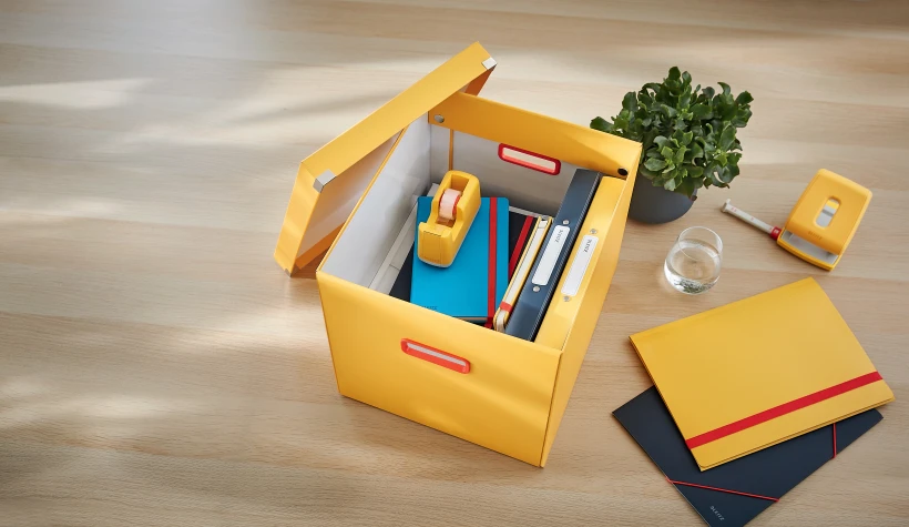 żółte pudełko z otwartą pokrywą i umieszczonymi wewnątrz akcesoriami, leżące na biurku 